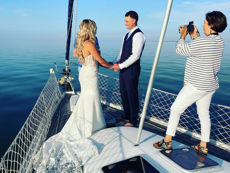 Bride and groom hold hands on a sailboat deck as a person photographs them with a phone.
