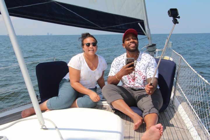 Two people on a sailboat taking a selfie with a selfie stick on a sunny day.