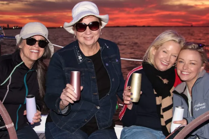 Four women smiling on a boat holding drinks at sunset.