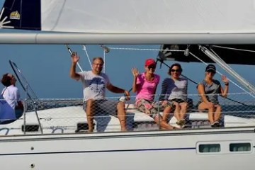Four people wave from a sailboat on a sunny day.
