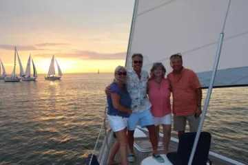 Four people on a sailboat smiling at sunset with other boats in the background.
