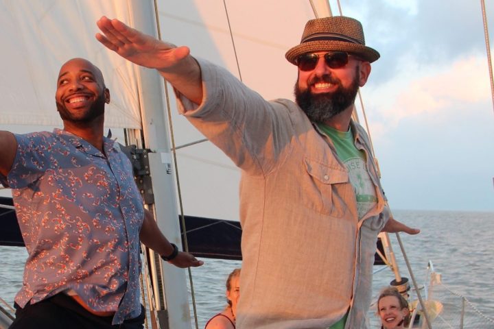 Two men striking playful poses on a sailboat at sea, with a woman sitting in the background.