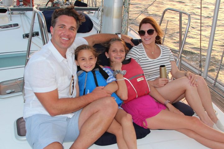 Family of four smiling on a sailboat deck at sunset.