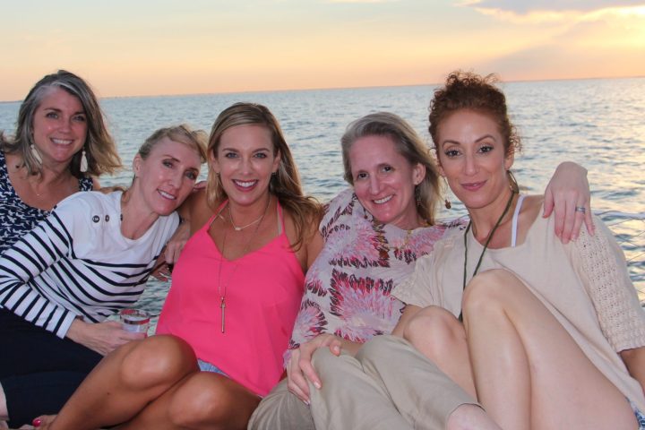 Five women posing on a boat at sunset, with water and city skyline in the background.