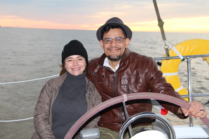 Two people smiling on a boat at sunset, one steering and both wearing jackets and hats.