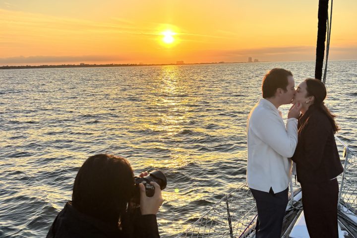 Couple kissing on a boat at sunset while a photographer captures the moment.