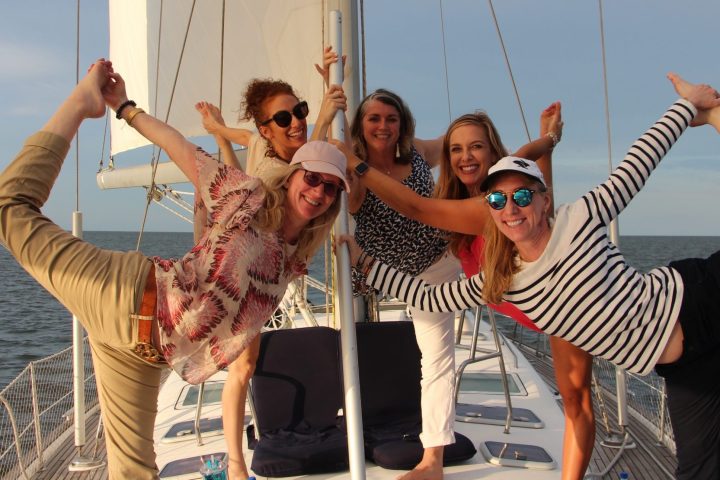 Five women posing energetically on a sailboat, with the ocean in the background.