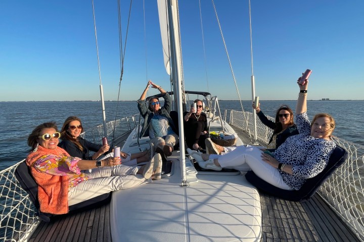Six people relaxing on a sailboat deck, holding drinks, with a clear blue sky and sea in the background.