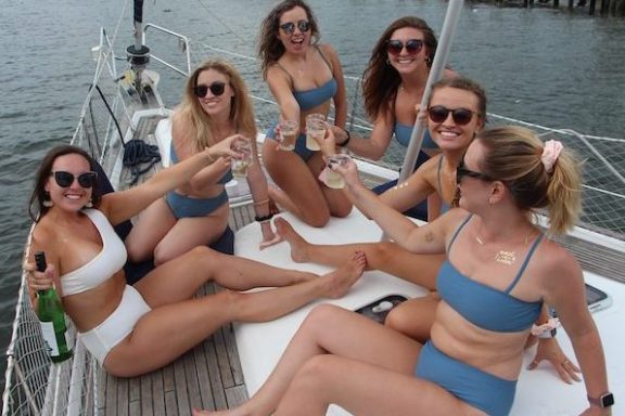Six women in swimsuits toasting drinks on a sailboat on a sunny day.