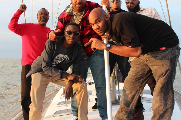 Six men posing on a sailboat with ocean and cloudy sky in the background.
