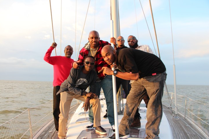 Six men posing on a sailboat with ocean and cloudy sky in the background.