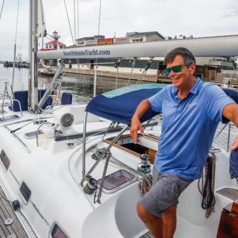 Man in blue shirt and sunglasses standing on sailboat docked near waterfront.