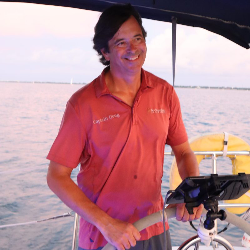 Smiling man steering a boat on a calm ocean, wearing a red shirt under a blue canopy.