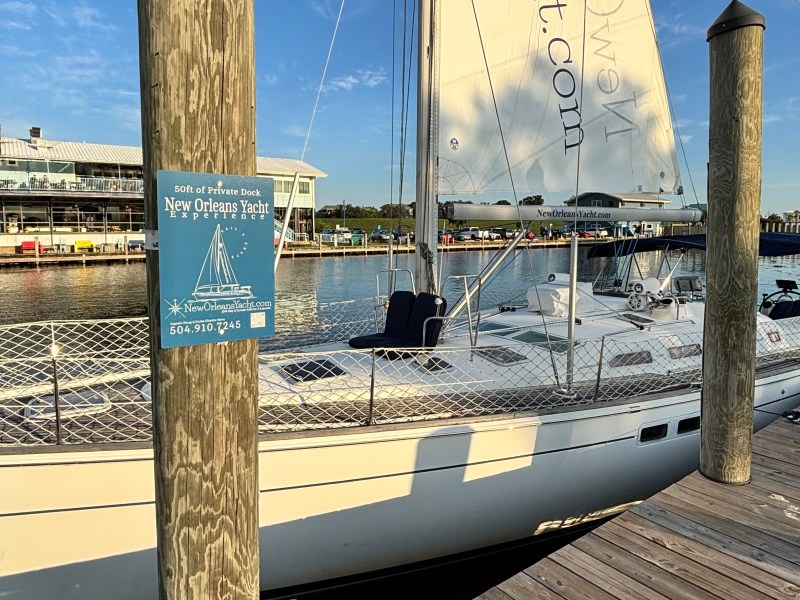 Docked sailboat with visible sail, against a backdrop of water and buildings, wooden poles in foreground.