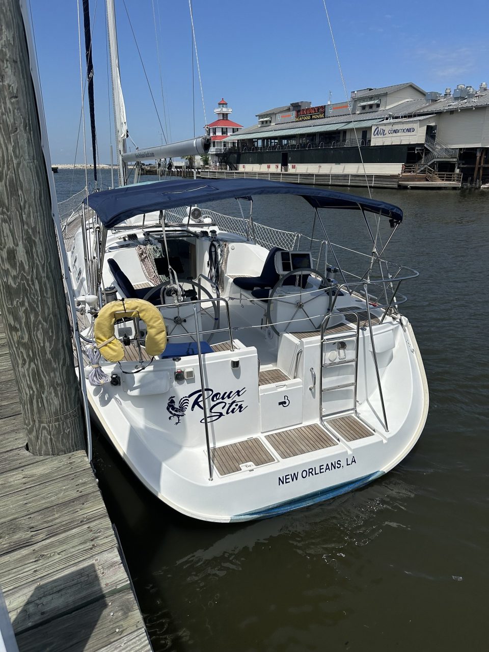 Sailboat docked by a pier with a restaurant and lighthouse in the background under a clear sky.