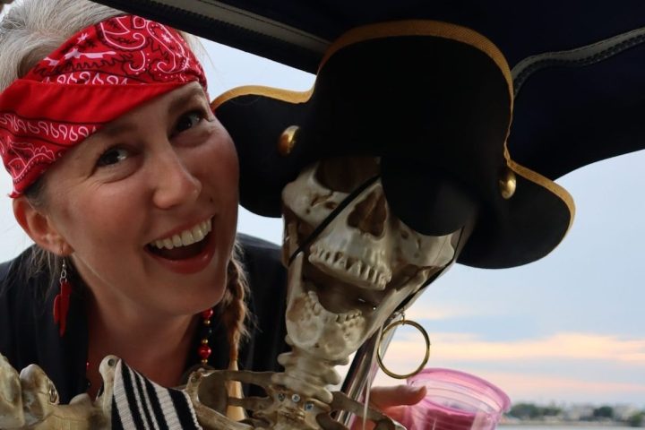 Woman in pirate outfit poses with skeleton on a boat.