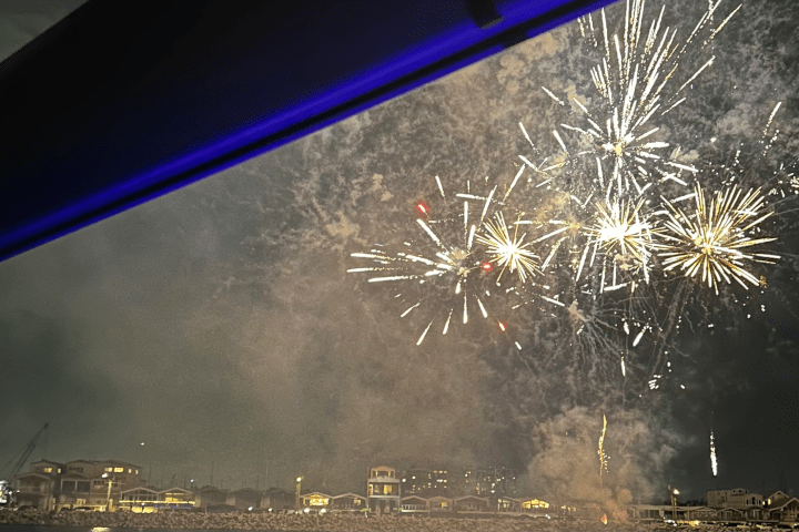 Fireworks burst in the night sky above a town with buildings and a thick cloud of smoke.