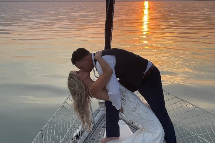 Couple kissing on sailboat at sunset, bride in gown and groom in suit.