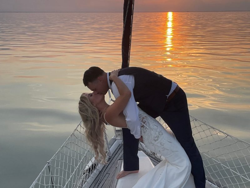 Couple kissing on sailboat at sunset, bride in gown and groom in suit.