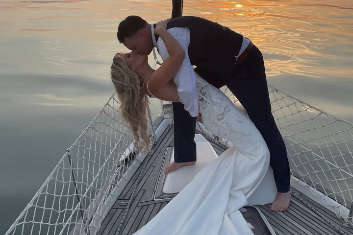 Couple kissing on sailboat at sunset, bride in gown and groom in suit.