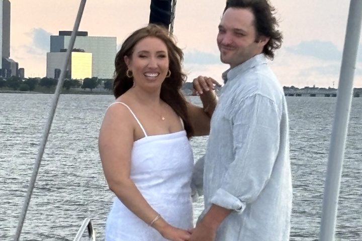 Couple on a sailboat at sunset, dressed in white, holding hands.