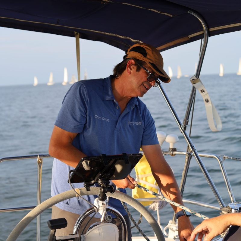 Man steering sailboat at sea, wearing hat and blue shirt, with other sailboats in background.