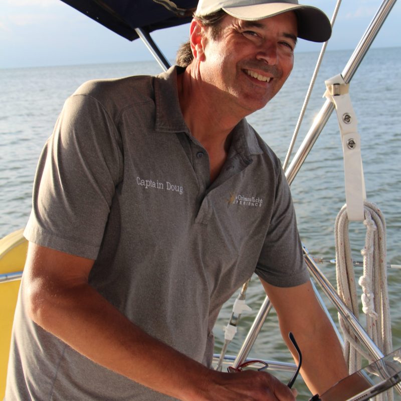 Smiling man at helm of sailboat, wearing a grey cap and shirt.