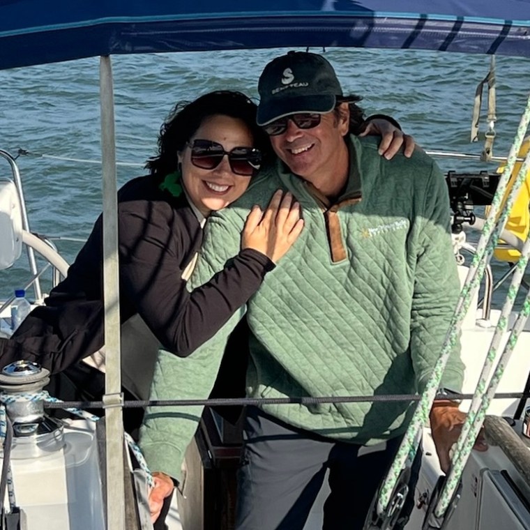 Two people smiling on a sailboat, wearing caps and sunglasses, with water in the background.