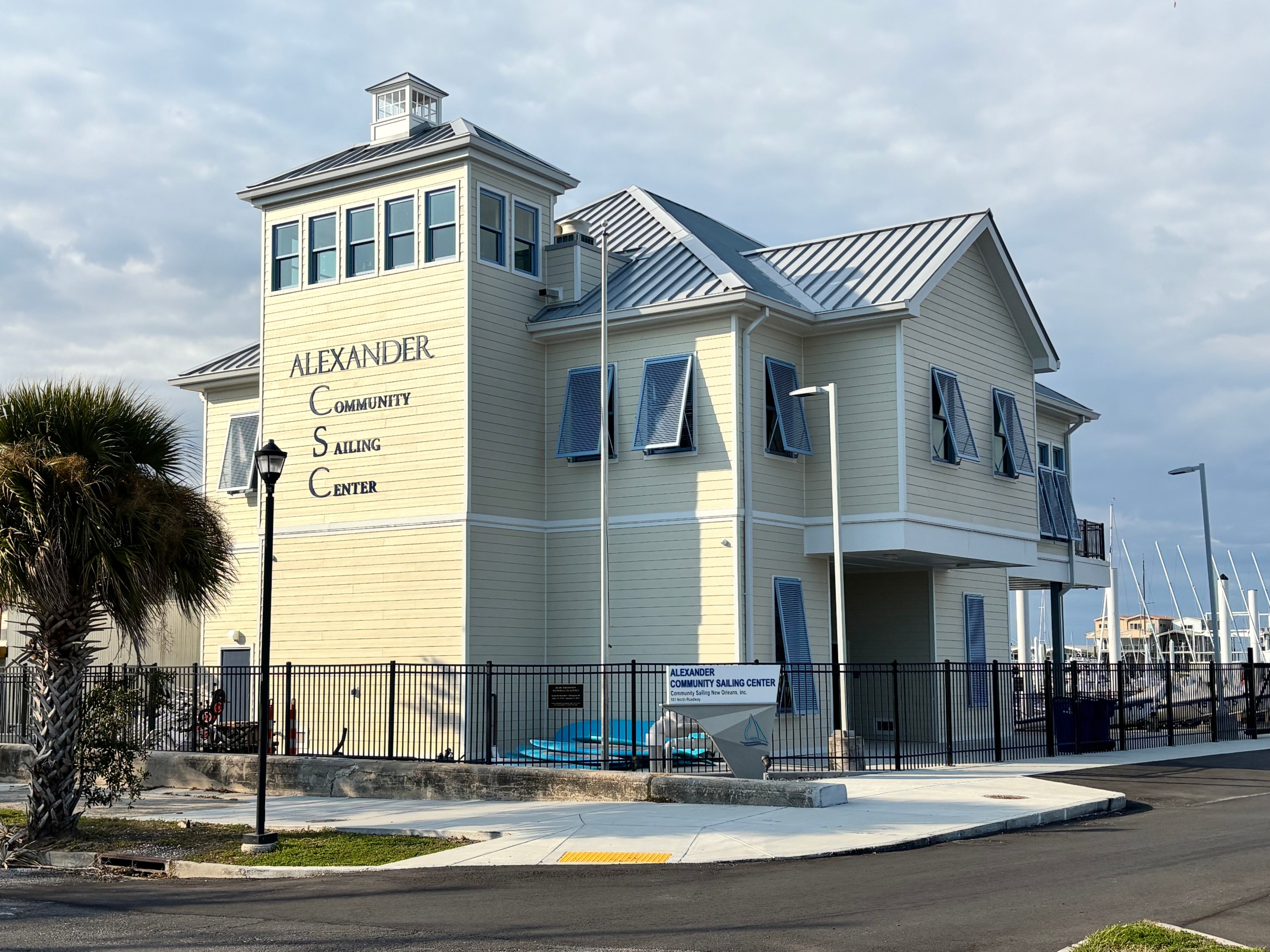 Yellow building with A-frame roof labeled 'Alexander Community Sailing Center' near palm trees and marina.